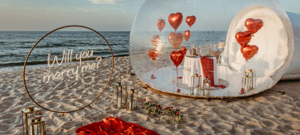 Sceneria zaręczyn na plaży nad morzem -  napis 'Will you marry me?' na piasku, czrwone balony w kształcie serc, płatki róż rozrzucone na piasku i romantyczna dekoracja na tle zachodzącego słońca.